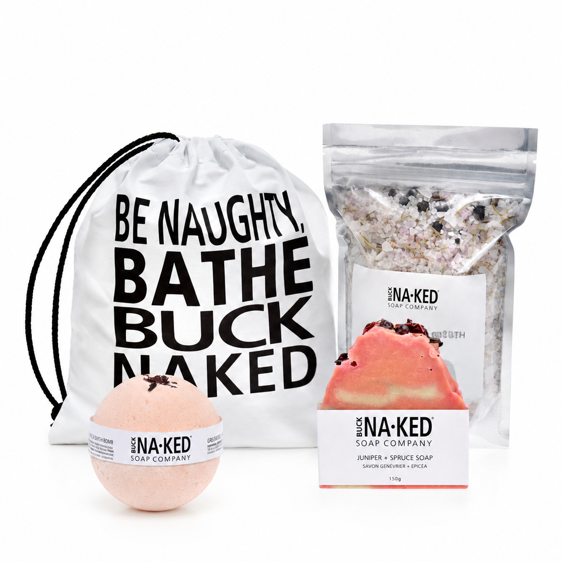 NA-KED Soap Company bath gift set featuring a white drawstring bag with black text, a peach-colored bath bomb, and a resealable pouch of bath salts on a white background.